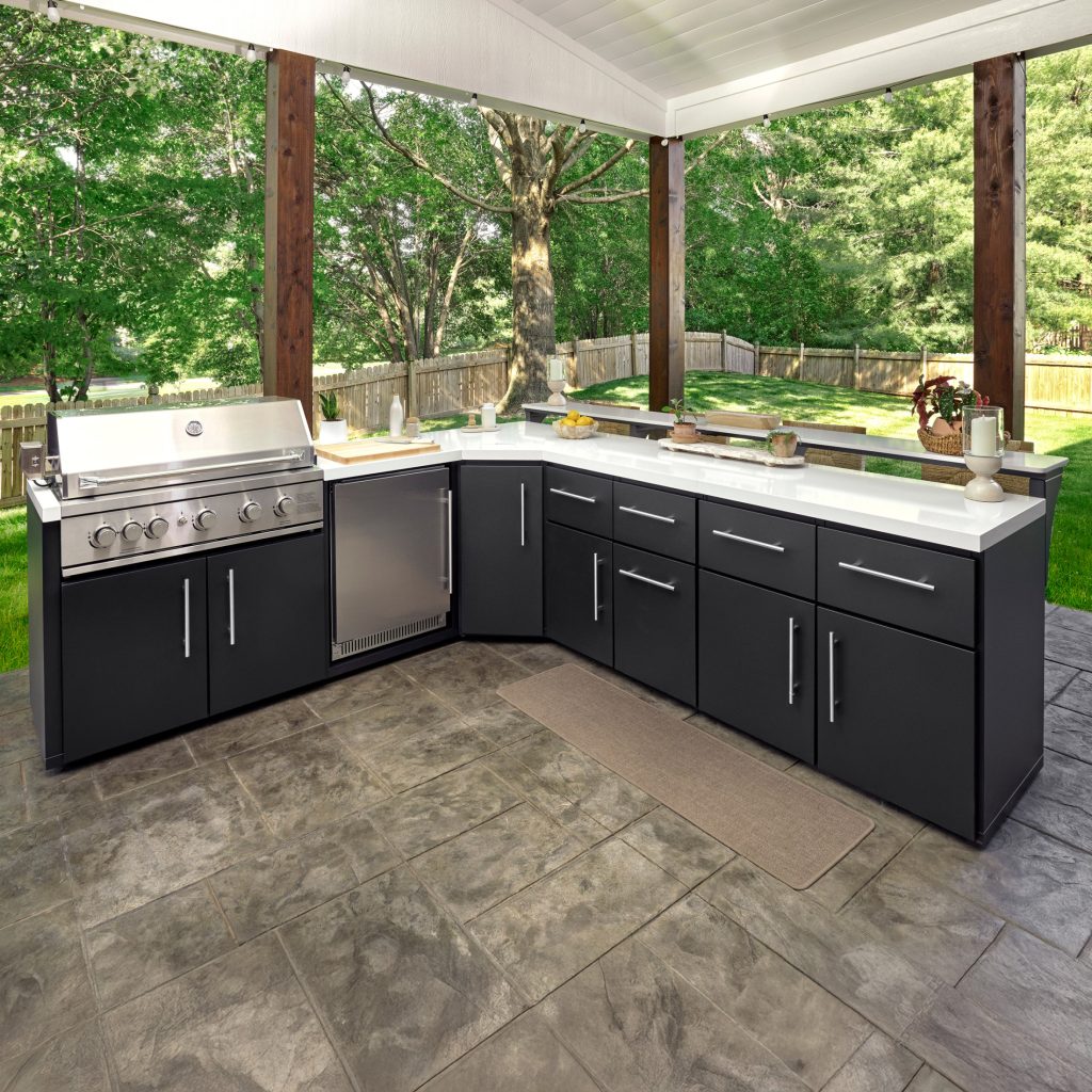 Large L-shaped outdoor kitchen island with black cabinetry, white countertops, stainless steel grill, and a raised bar top under a covered patio.