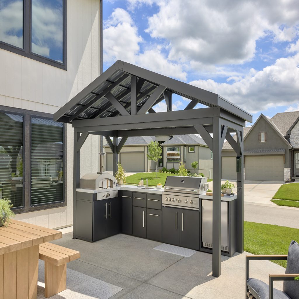 Lancaster black steel pavilion featuring an L-shaped outdoor kitchen with a pizza oven, gas grill, and built-in refrigerator on a concrete patio.