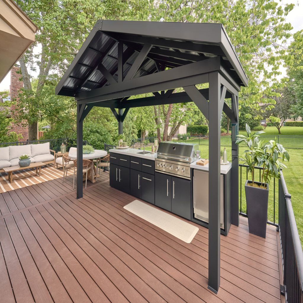 Lancaster linear outdoor kitchen with a professional gas grill and built-in refrigerator installed on a raised wood deck under a black steel pavilion.