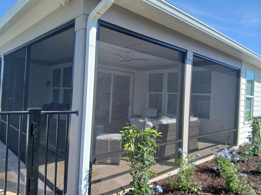 Exterior view showing the expert craftsmanship of the screen room installation in Nexton, SC. The durable dark framing contrasts sharply with the house and is finished with beautiful local landscaping and blue flowers.